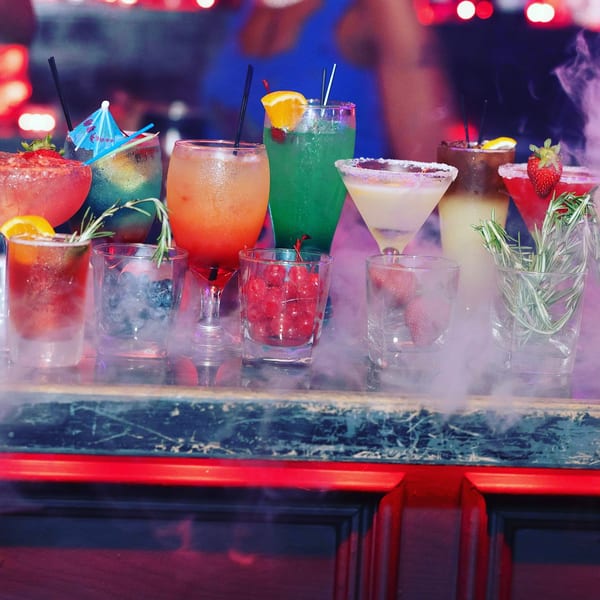 A bar top displays a variety of colorful cocktails, some garnished with fruit and herbs, surrounded by smoke.
