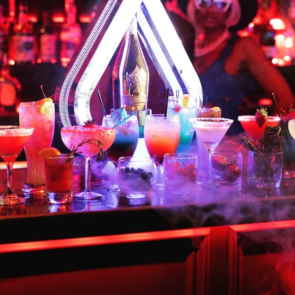 A bar counter with many colorful cocktails and a bartender in the background under red lighting.