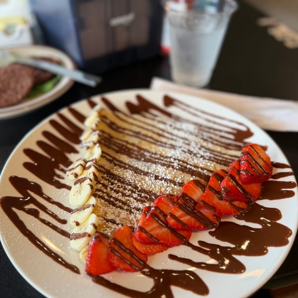 A sweet crepe drizzled with chocolate, powdered sugar, sliced bananas, and strawberries on a white plate.