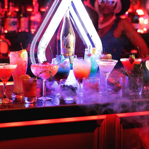 A bar counter with a variety of colorful cocktails and a bottle of champagne in a neon-lit setting.