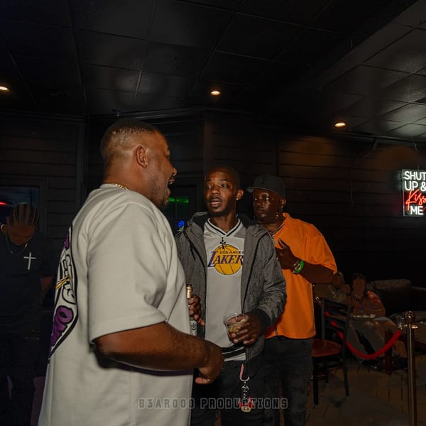 Three men conversing in a dimly lit bar with neon signs and a DJ booth in the background.